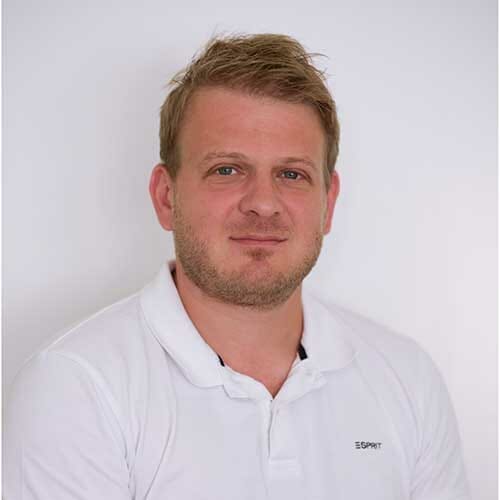 Mann mit kurzem blondem Haar und gestutztem Bart in weißem Hemd vor hellem Hintergrund.