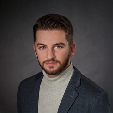 Junger Mann mit kurzen braunen Haaren und Bart in grauem Rollkragenpullover, dunkler Blazer, schlichter Hintergrund.