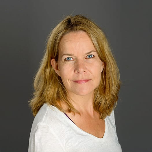 Blonde Frau mit blauen Augen in weißem Oberteil posiert vor grauem Hintergrund, neutraler Ausdruck.