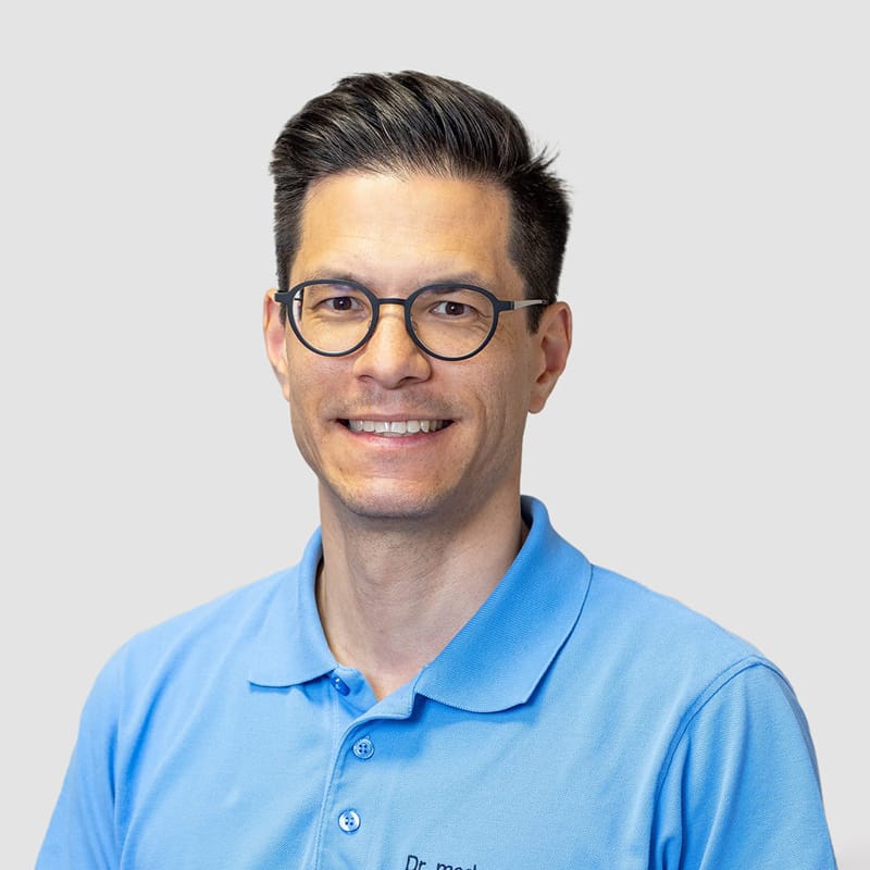 Lächelnder Mann mit kurzen dunklen Haaren und Brille in einem hellblauen Poloshirt.