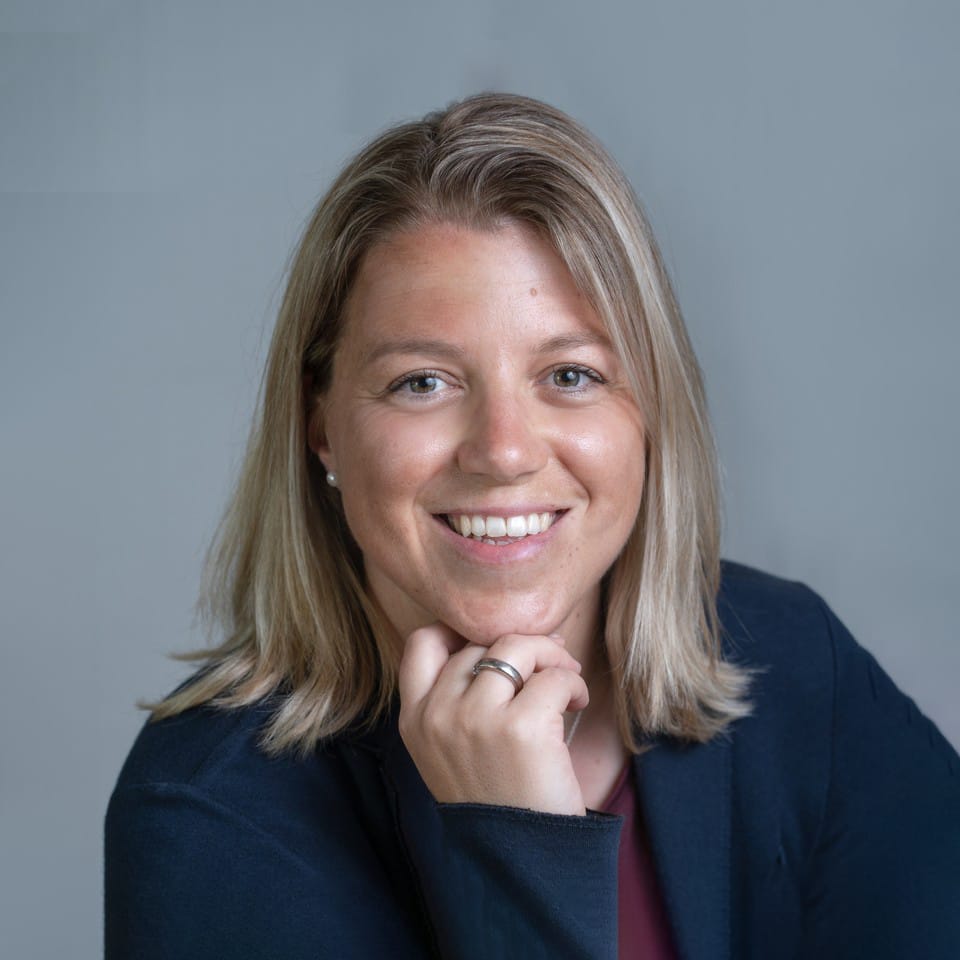 Frau mit glattem blondem Haar, lächelnd, Hand am Kinn, dunkler Blazer, grauer Hintergrund.