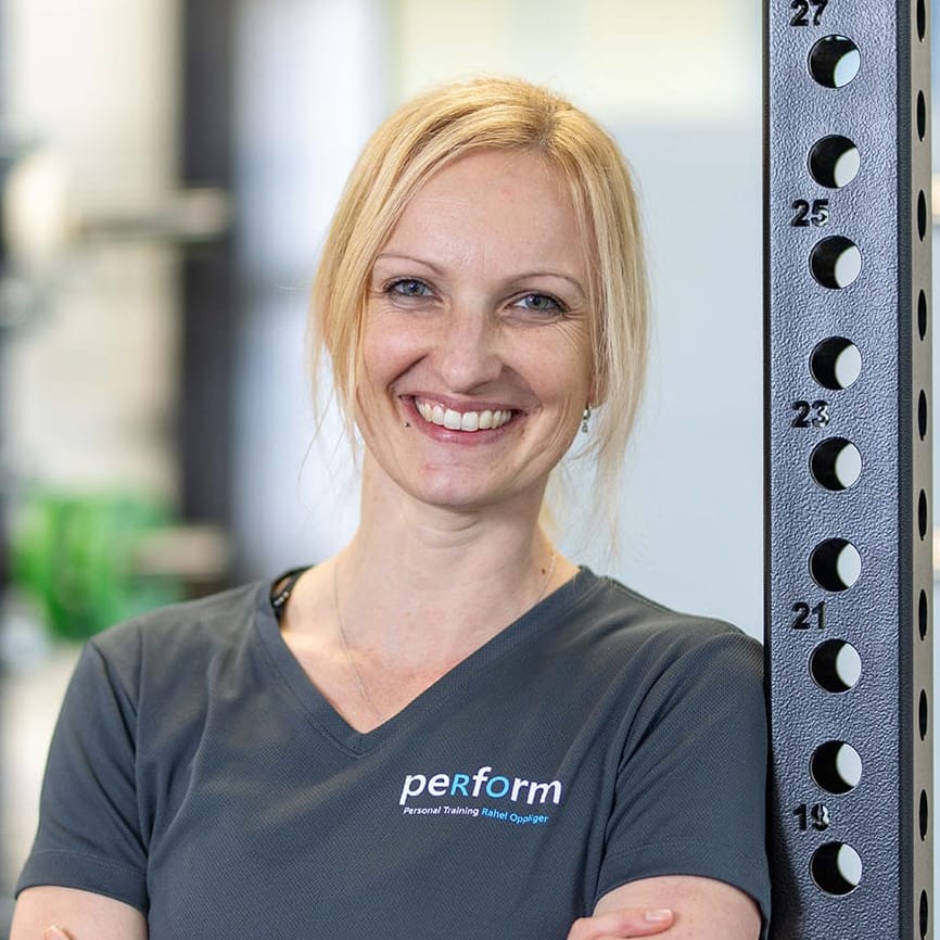 Eine lächelnde blonde Frau im grauen "Perform"-Shirt steht mit verschränkten Armen in einem Fitnessstudio.