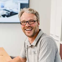 Ein lächelnder Mann mit grauem Haar, Brille und Bart sitzt an einem Tisch in einem hellen Raum.