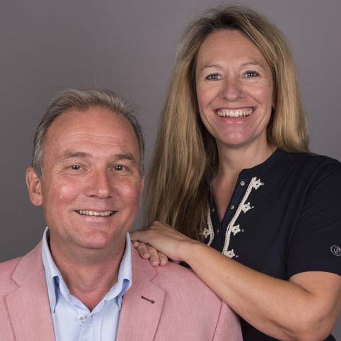 Ein lächelnder Mann im rosa Blazer sitzt, während eine blonde Frau hinter ihm steht, beide auf grauem Hintergrund.