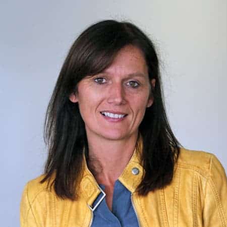 Lächelnde Frau mit glattem braunem Haar in gelber Jacke und blauem Oberteil, einfarbiger Hintergrund.