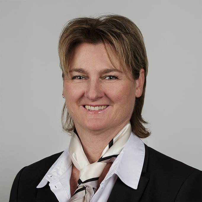 Frau mit hellbraunem Haar, schwarzem Blazer, weißem Hemd, Schal, lächelt in die Kamera.