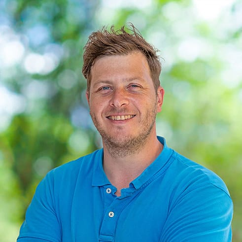 Lächelnder Mann mit kurzen braunen Haaren und Bart in blauem Poloshirt im Freien, unscharfe Grünfläche.