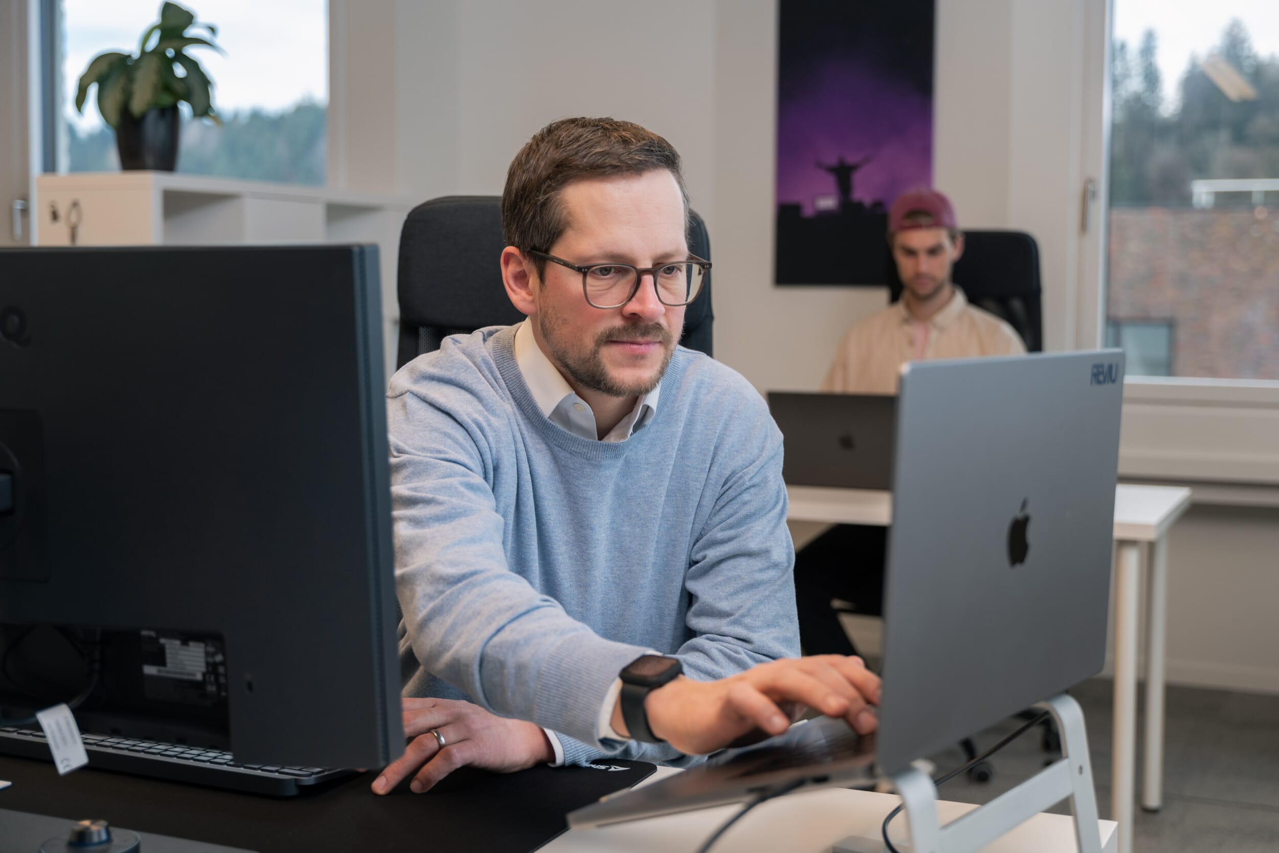 Zwei Männer arbeiten in einem modernen Büro an Computern; einer trägt eine Brille und eine Smartwatch.