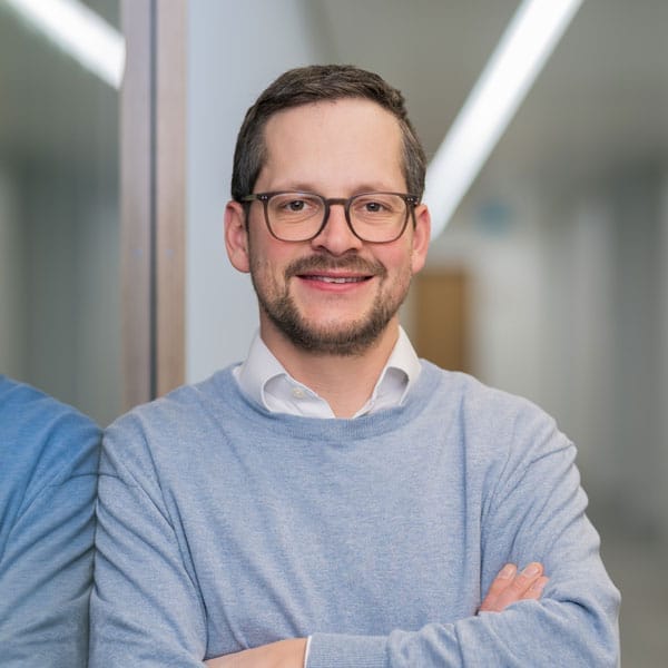 Mann mit kurzen dunklen Haaren, Brille und Bart lächelt mit verschränkten Armen in einem Haus.