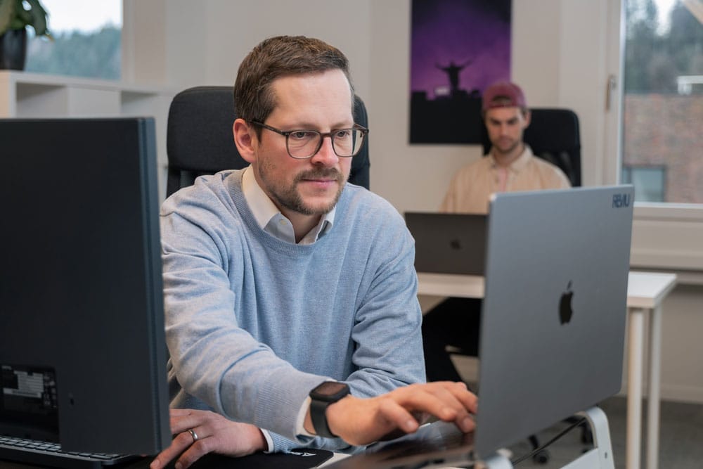 Mann mit Brille und Bart arbeitet in einem Büro am Laptop, eine weitere Person im Hintergrund.