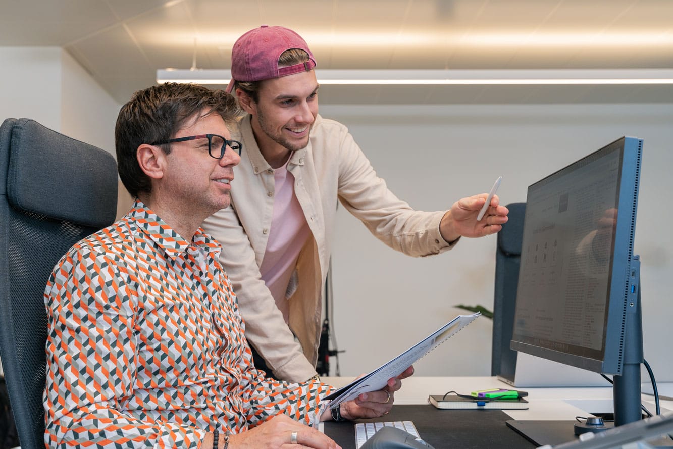 Zwei Männer arbeiten an einem Schreibtisch, einer sitzt mit Papieren, der andere zeigt auf einen Computer.
