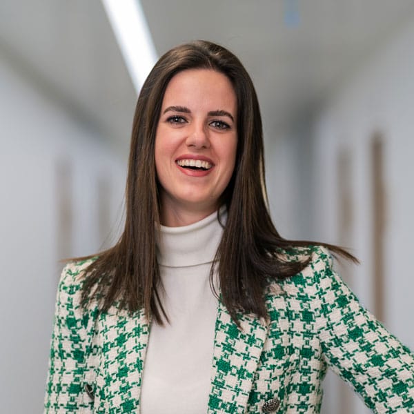 Frau mit glattem braunem Haar und grünem Blazer lächelt in einem hellen, modernen Flur.