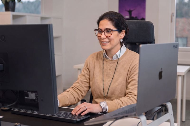 Eine Frau mit Brille und beigem Pullover arbeitet an einem Schreibtisch mit einem Computer und einem Laptop.
