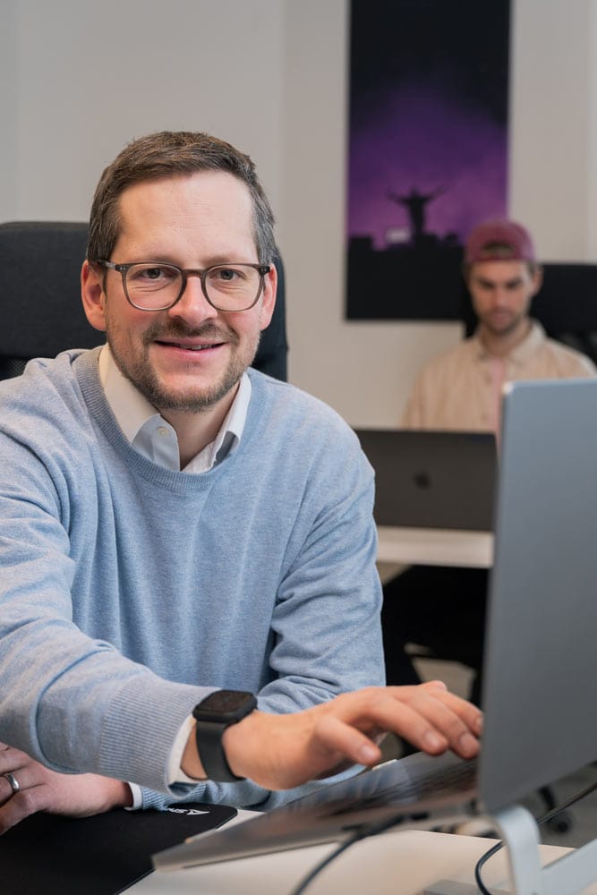 Man arbeitet mit Brille und blauem Pullover lächelnd am Laptop in modernem Büro.