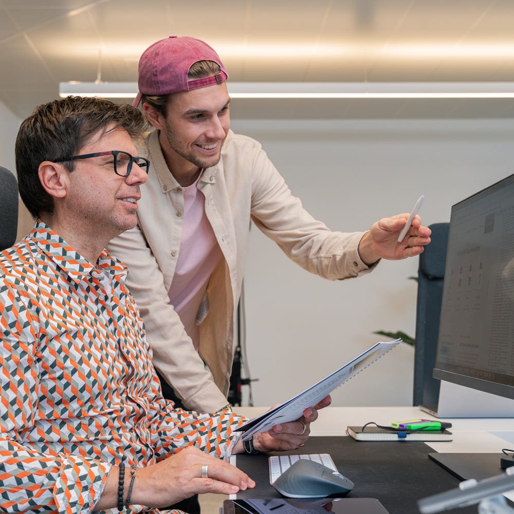 Zwei Männer in einem Büro, einer sitzend am Computer, der andere stehend und auf einen Monitor zeigend.