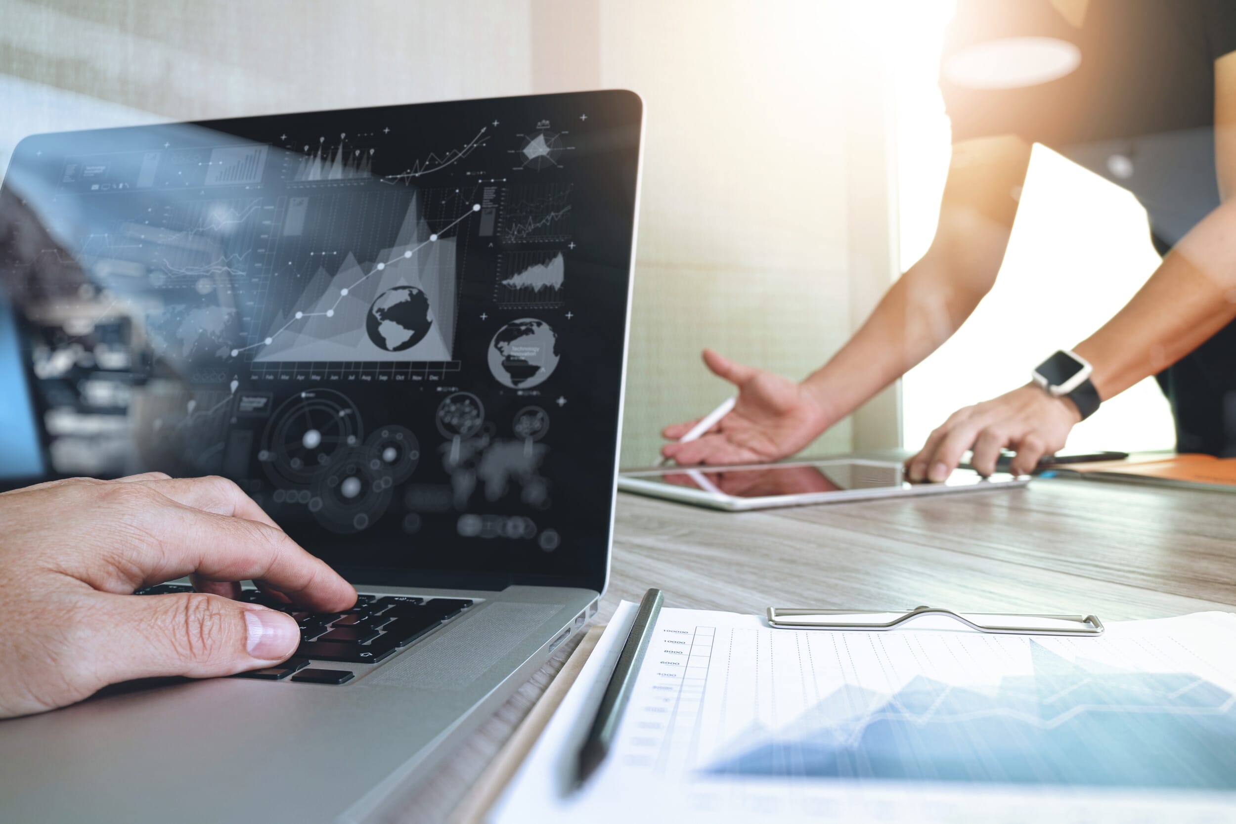 Eine Person tippt auf einem Laptop mit Diagrammen, eine andere benutzt ein Tablet. Diagramme auf einem sonnenbeschienenen Tisch.