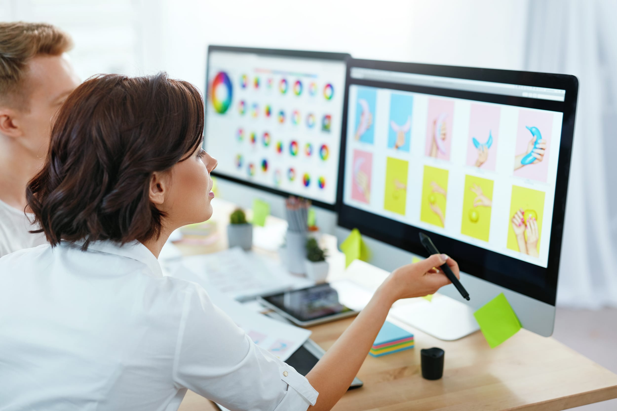 Frau mit Stift an einem Schreibtisch mit zwei Monitoren, auf denen bunte grafische Designs angezeigt werden.