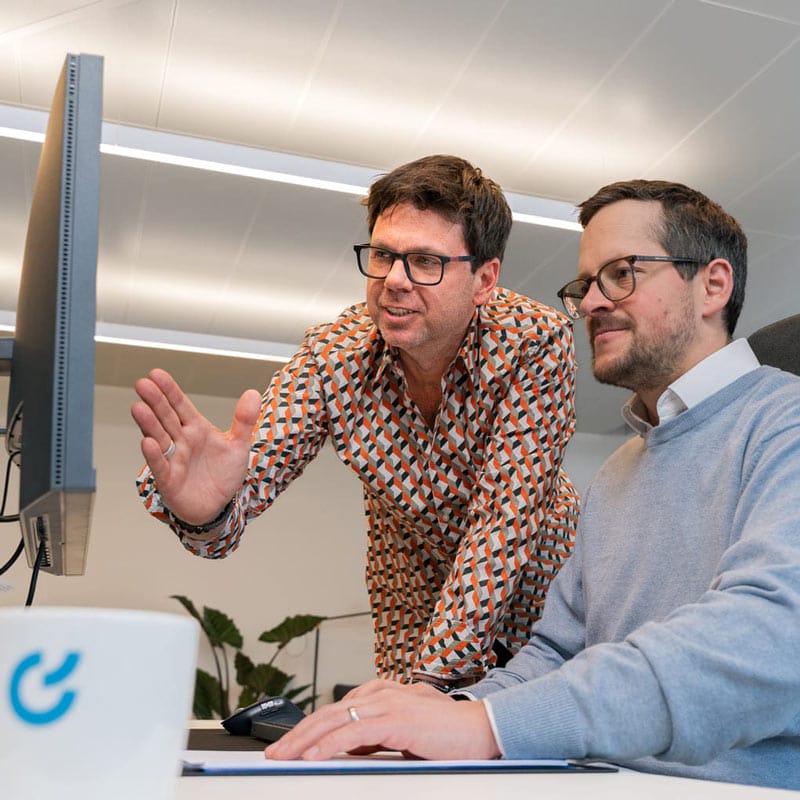 Zwei Männer in Geschäftskleidung arbeiten gemeinsam an einem Computer und besprechen, was auf dem Bildschirm zu sehen ist.