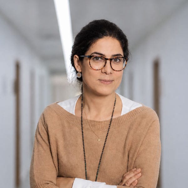 Eine Frau mit dunklem Haar und Brille steht im Flur, die Arme verschränkt, einen hellbraunen Pullover über einem weißen Hemd.