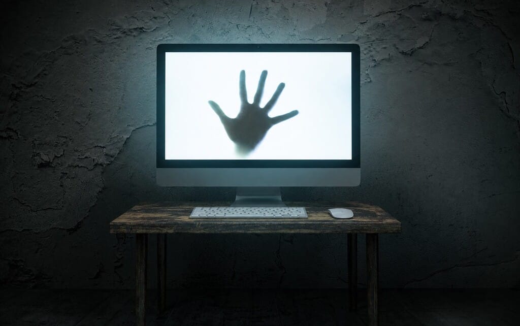 Computer mit einer schattenhaften Hand auf dem Bildschirm auf einem Holzschreibtisch vor einer dunklen Wand.