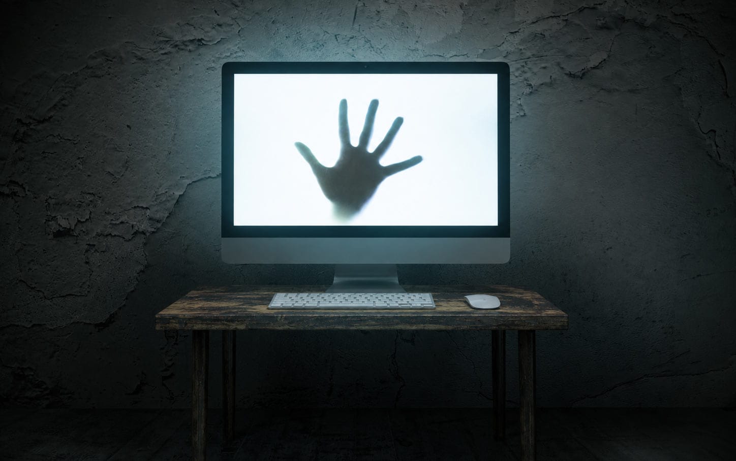 Computer mit einer schattenhaften Hand auf dem Bildschirm auf einem Holzschreibtisch vor einer dunklen Wand.